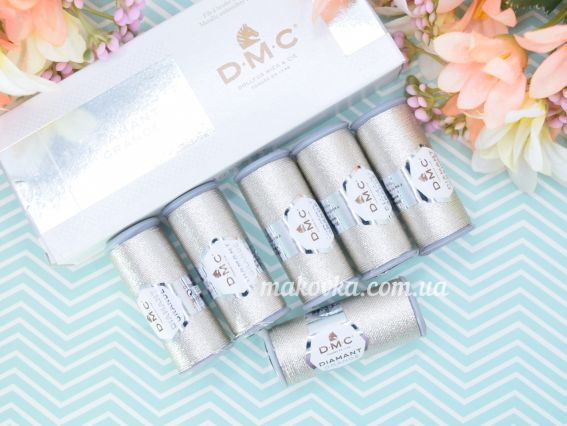 П'ять котушок металізованого муліне DMC Diamant Grande срібного кольору, викладених в один рівний ряд на фоні коробки та декоративних квітів.