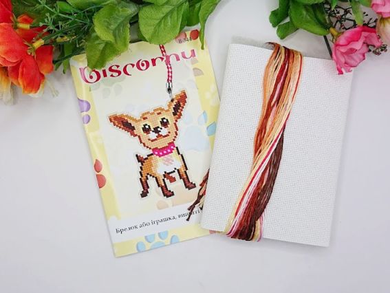 Вміст набору для вишивання брелока Biscornu: паперова обкладинка з малюнком чихуахуа та комплект кольорових ниток муліне на білому органайзері.