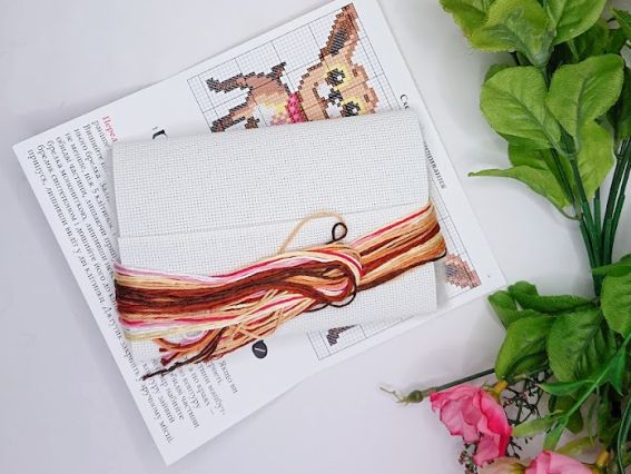 На білій пластиковій канві (спеціальна сітчаста основа для вишивання) лежить пасмо ниток муліне коричневих та рожевих відтінків. Поруч видно частину кольорової схеми та декоративні квіти. Фото демонструє основні матеріали для створення брелока.