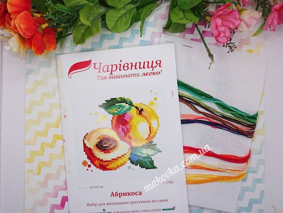 Обкладинка набору для вишивання «Абрикоса» (N-1705) від ТМ «Чарівниця» поруч із білою канвою та пасмом ниток муліне теплих помаранчевих, червоних та зелених кольорів.