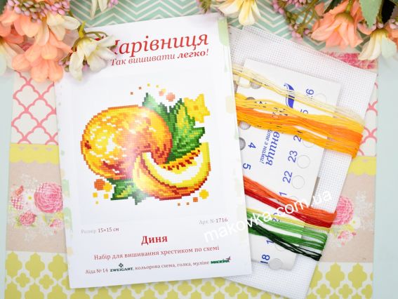 Обкладинка набору для вишивання N1716 Диня від ТМ «Чарівниця» поруч із білою канвою та пасмом ниток муліне теплих помаранчевих, червоних та зелених кольорів.