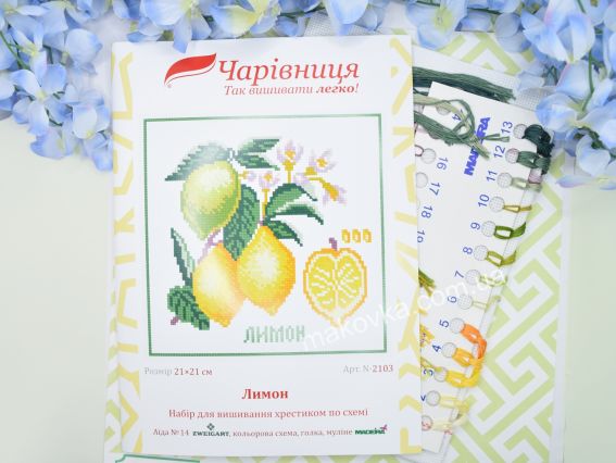 Обкладинка набору для вишивання N2103 Лимон від ТМ «Чарівниця» поруч із пасмом ниток муліне жовтих, оранжевих та зелених кольорів на блакитному тлі з квітами.