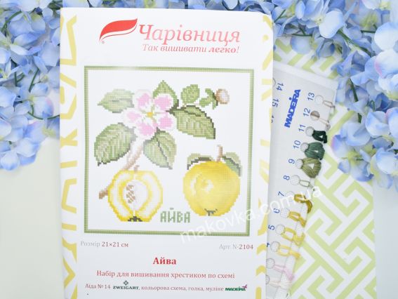 Обкладинка набору для вишивання N2104 Айва від ТМ «Чарівниця» поруч із пасмом ниток муліне жовтих, пісочних та зелених кольорів на блакитному тлі з квітами.